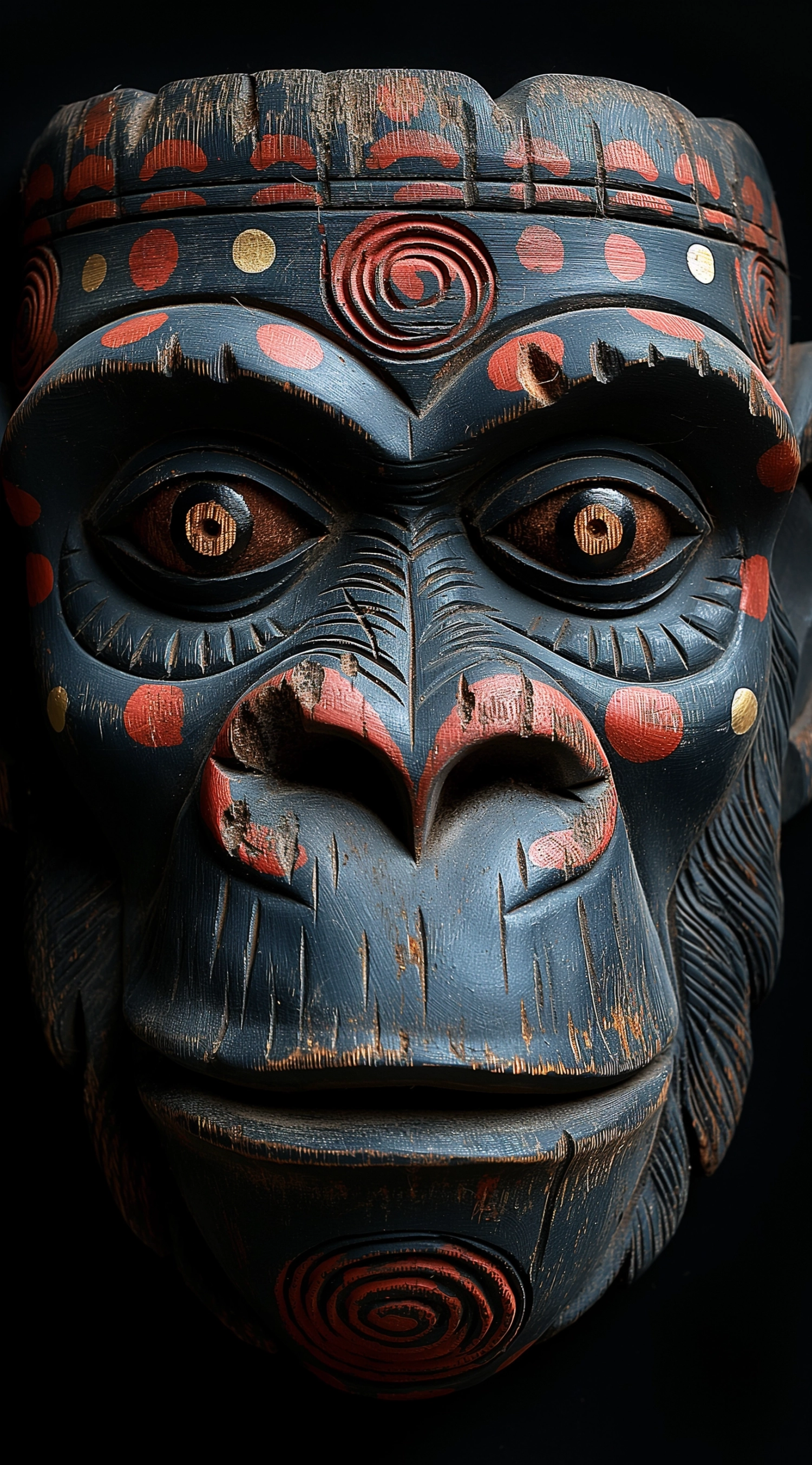 black background, top view of an angry monkey mask made from dark wood with painted eyes and mouth open holding out its hand to you. the wooden carved features on it have been decorated in patterns by native art carving artistes, using red dots for lips, white stripes as eyebrows and grey or black lines for eyelashes. the overall look is eerie but also comical.