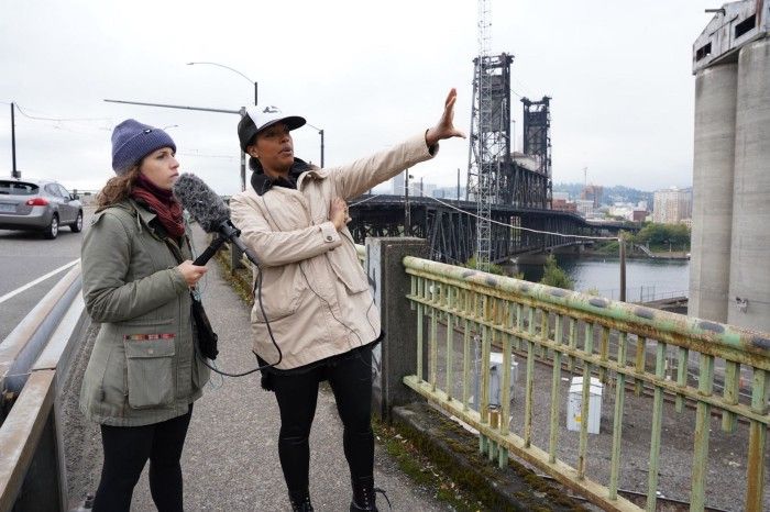 Photograph of two women, one holding a microphone, standing on a bridge's sidewalk, between the road and an industrial lot.