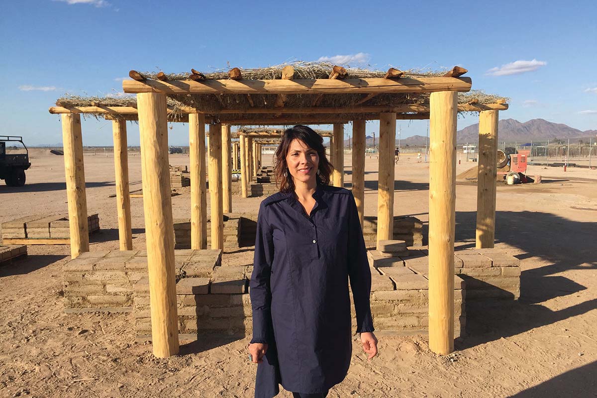 A woman stands in front of a wooden structure in the desert