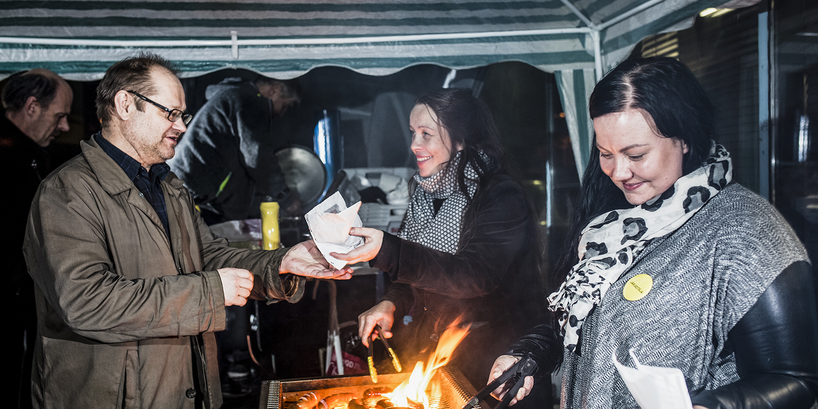 Kaksi naista paistaa makkaraa, toinen ojentaa miehelle yhden.