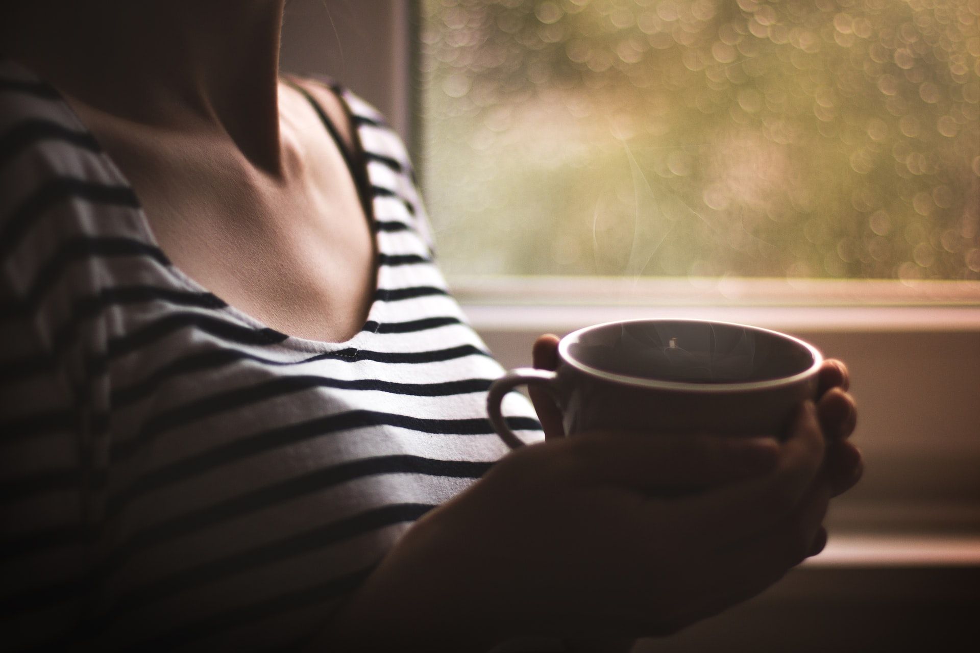 Nainen seisoo ikkunan edessä ja pitelee kahvikuppia