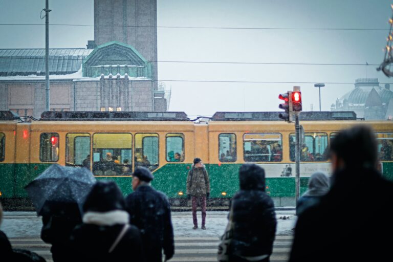 Mies seisoo llumipyryssä, rautatientorin edessä liikennevaloissa, raitiovaunupysäkin luona. Ihmisten silhuetteja näkyy.