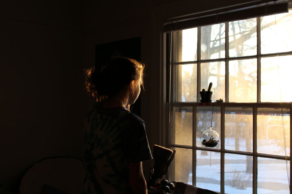 kuvassa hahmo katsoo pimeässä huoneessa ulkona paistavaan aurinkoiseen maisemaan