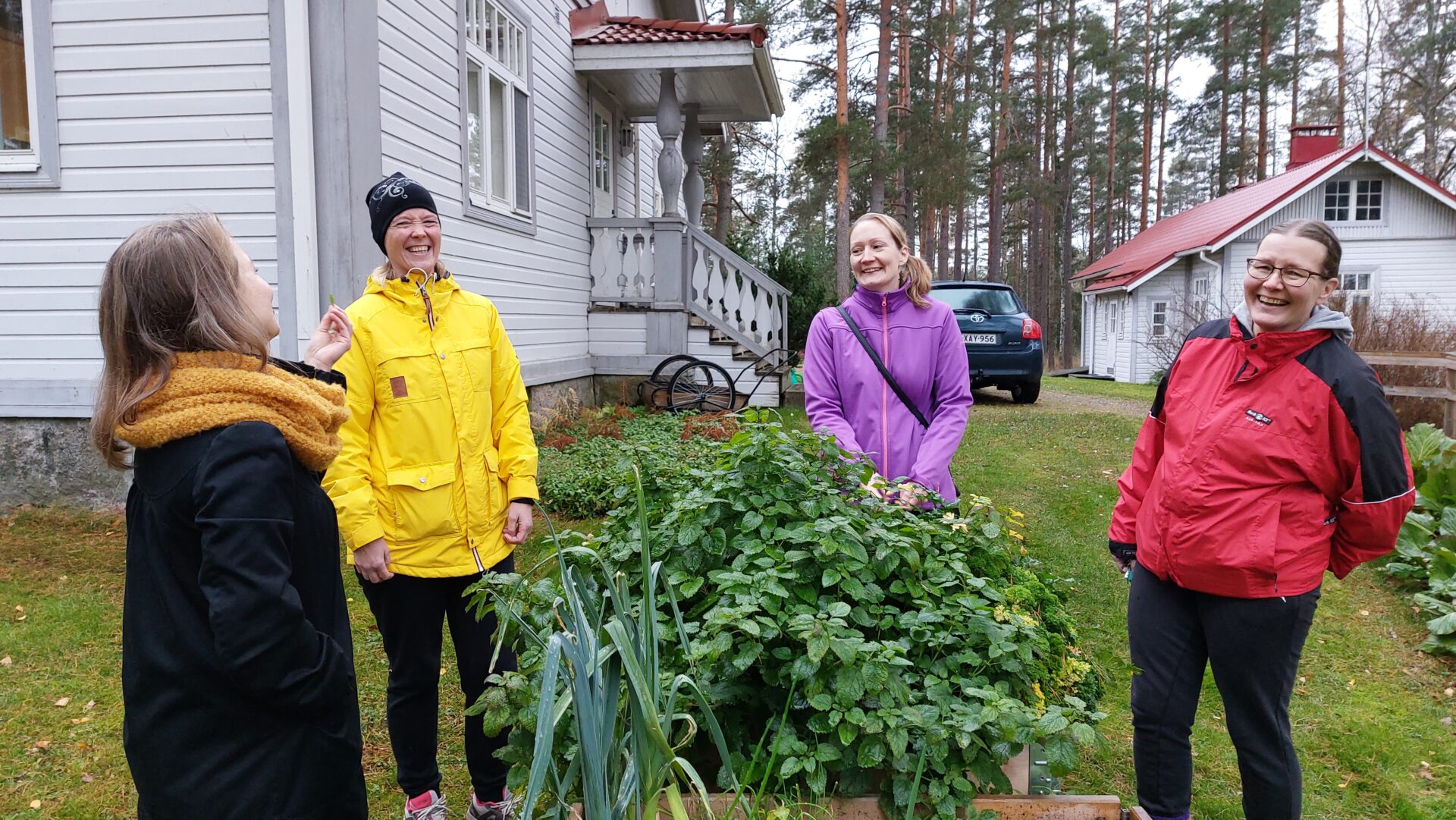 Neljä naista vanhan huvilan edessä seisovat ja nauravat viljelylaatikon äärellä.