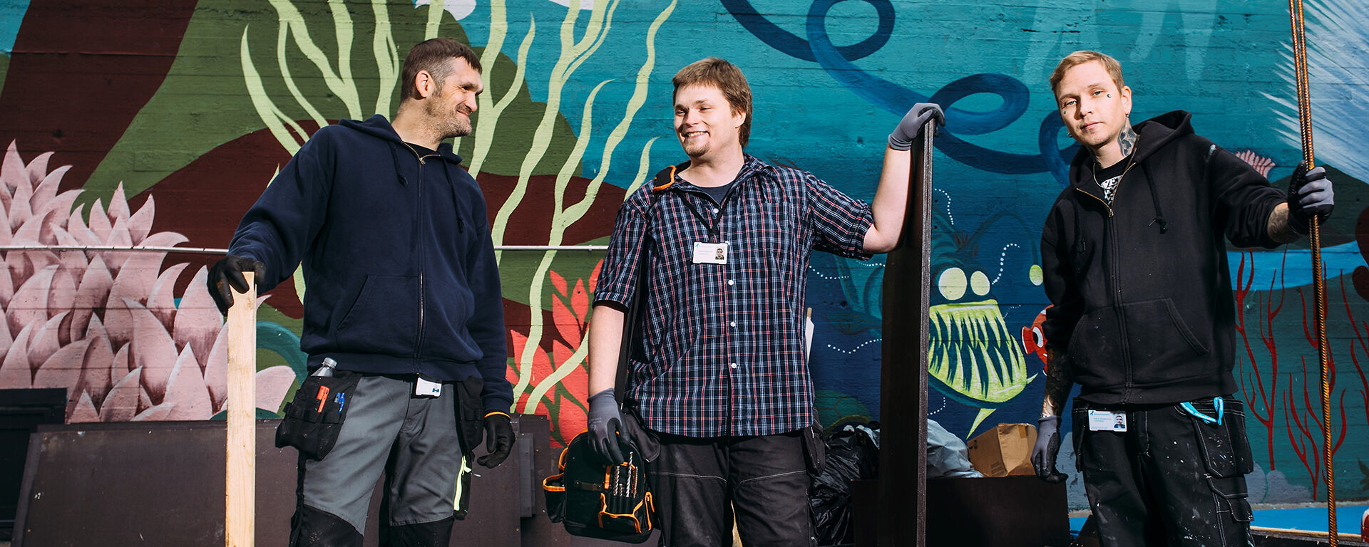Kolme iloista miestä graffitiseinän edessä työkalut kädessä
