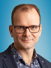 Siilitukkainen, silmälasipäinen mies, jolla on kauluspaita ja pikkutakki