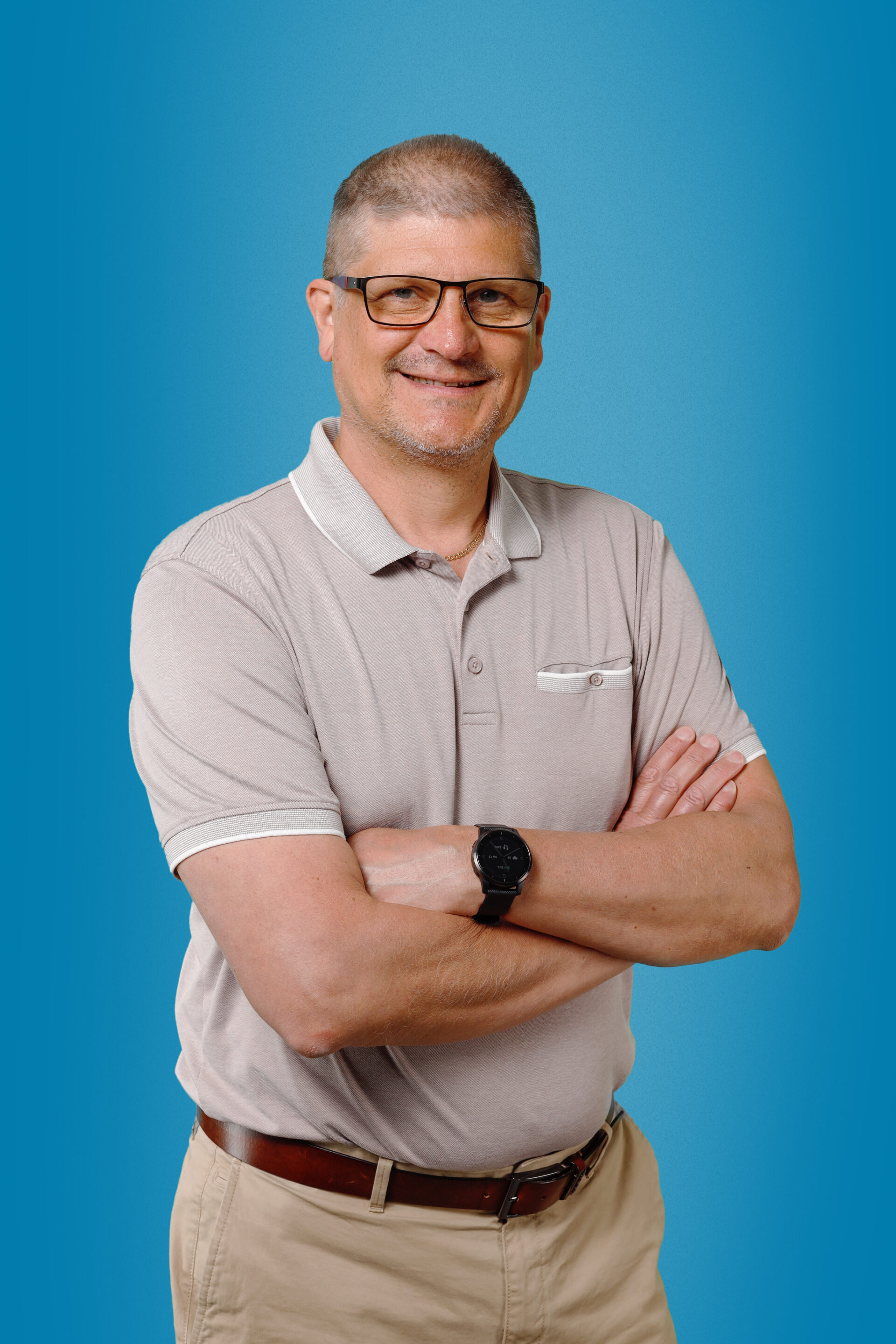 Hymyilevä, sänkitukkainen ja silmälasipäinen mies, jolla on käsivarret ristissä. Hänellä pikee-paita, jonka ylin nappi on auki, ja iso rannekello.