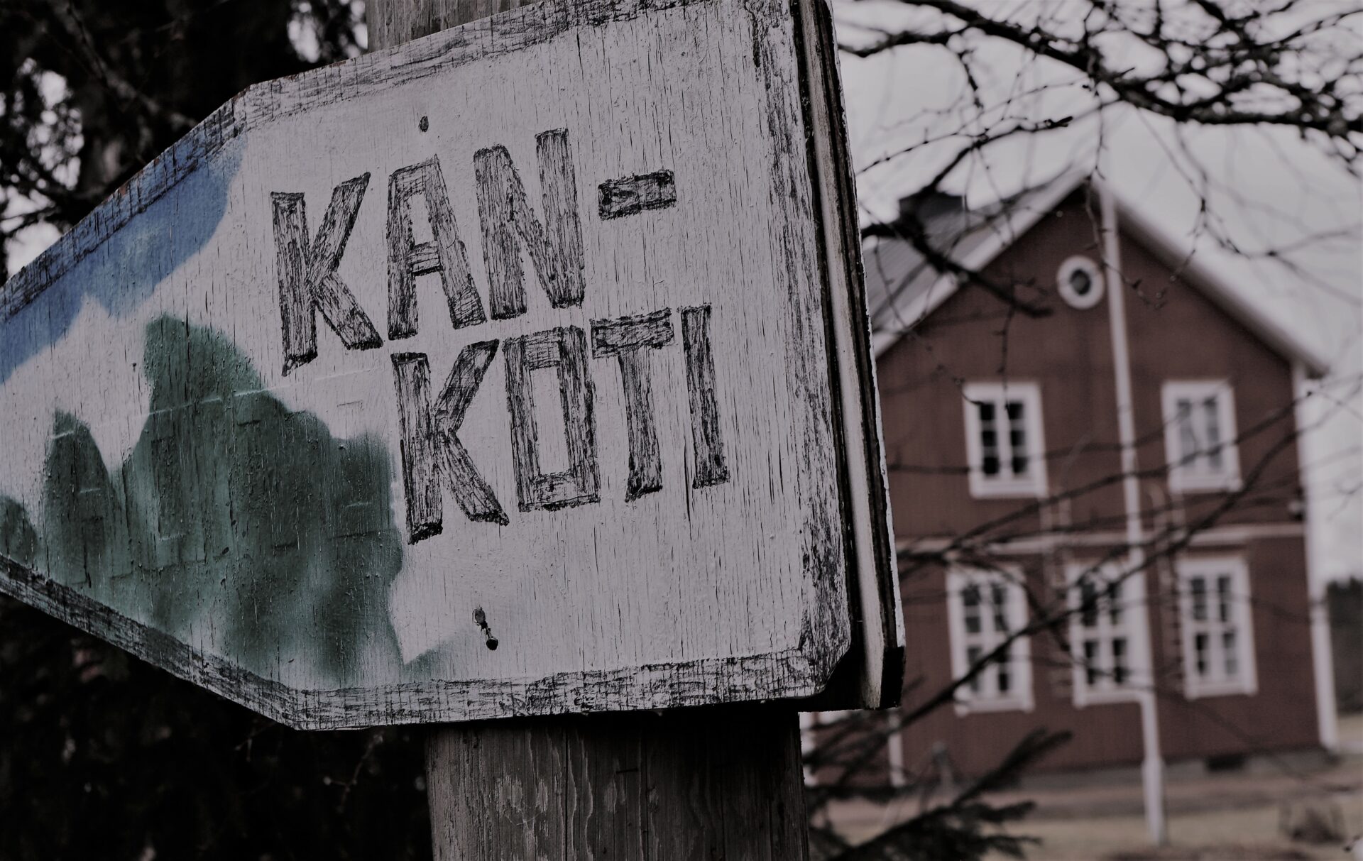 Etualalla puinen, hieman kömpelä nuolenmuotoinen viitta, jossa lukee KAN-koti. Taka-alalla kaksikerroksisen harjakattoisen puutalon pääty.