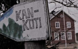 Etualalla puinen, hieman kömpelä nuolenmuotoinen viitta, jossa lukee KAN-koti. Taka-alalla kaksikerroksisen harjakattoisen puutalon pääty.