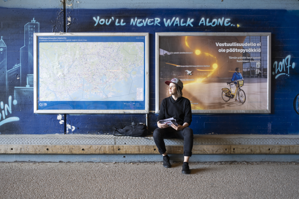 Nainen istuu katukivetyksellä, taustalla teksti "You'll never walk alone.", valokuva.