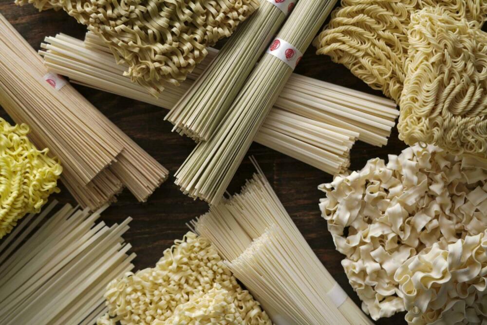 Assorted Asian noodles including flat noodles, ramen, knife cut noodles, and wheat noodles displayed on a wooden table.