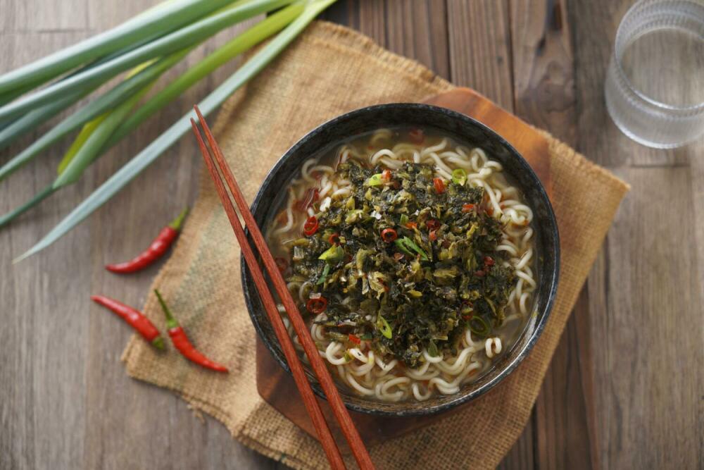 A cozy and flavorful bowl of SING-LIN FOODS beef flavored ramen topped with pickled mustard greens, scallions, and sliced chili peppers.