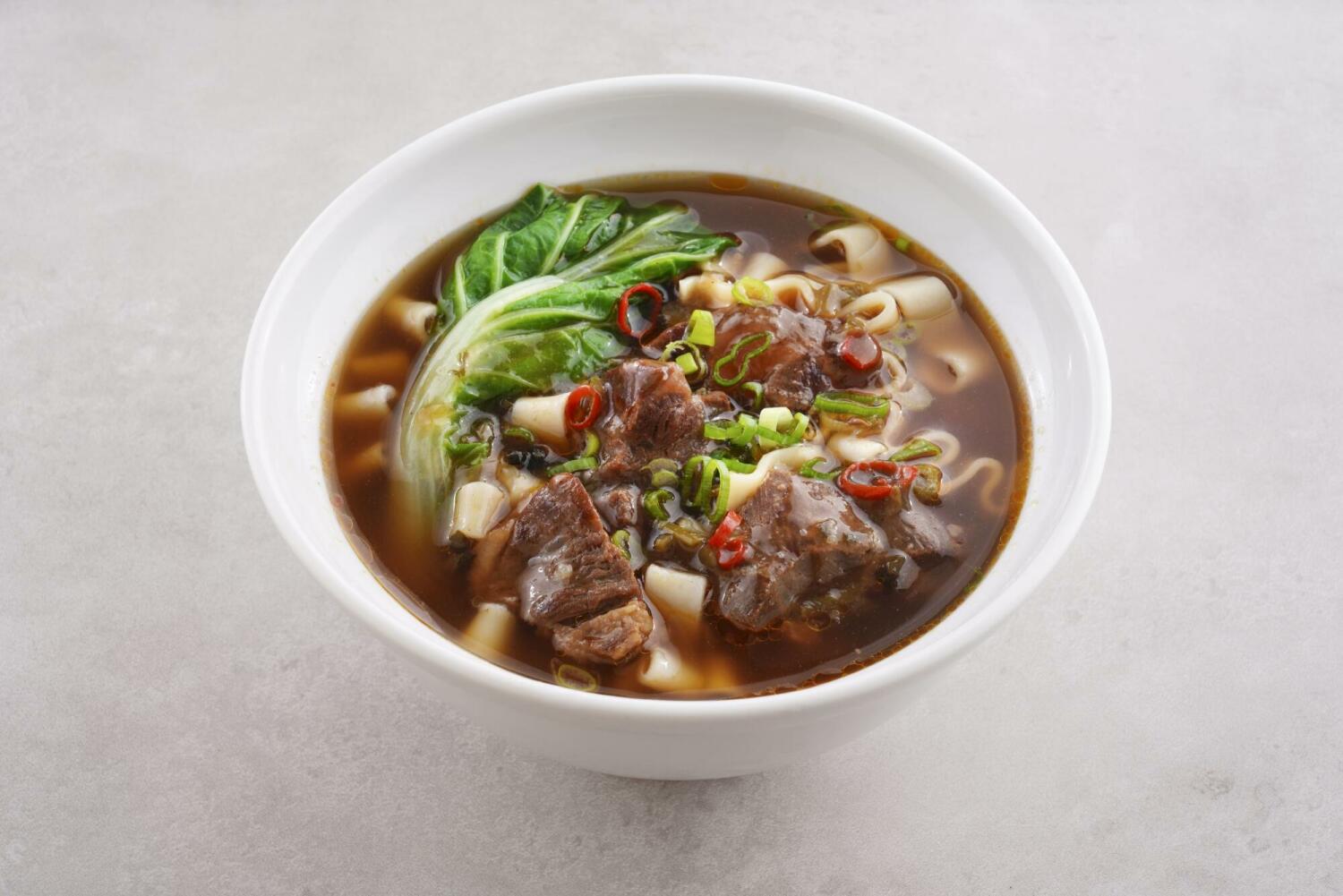 紅燒牛肉麵盛裝於白色碗中，濃郁湯頭搭配寬麵、牛肉、青菜與蔥花，呈現適合風味較為厚重的麵條料理。