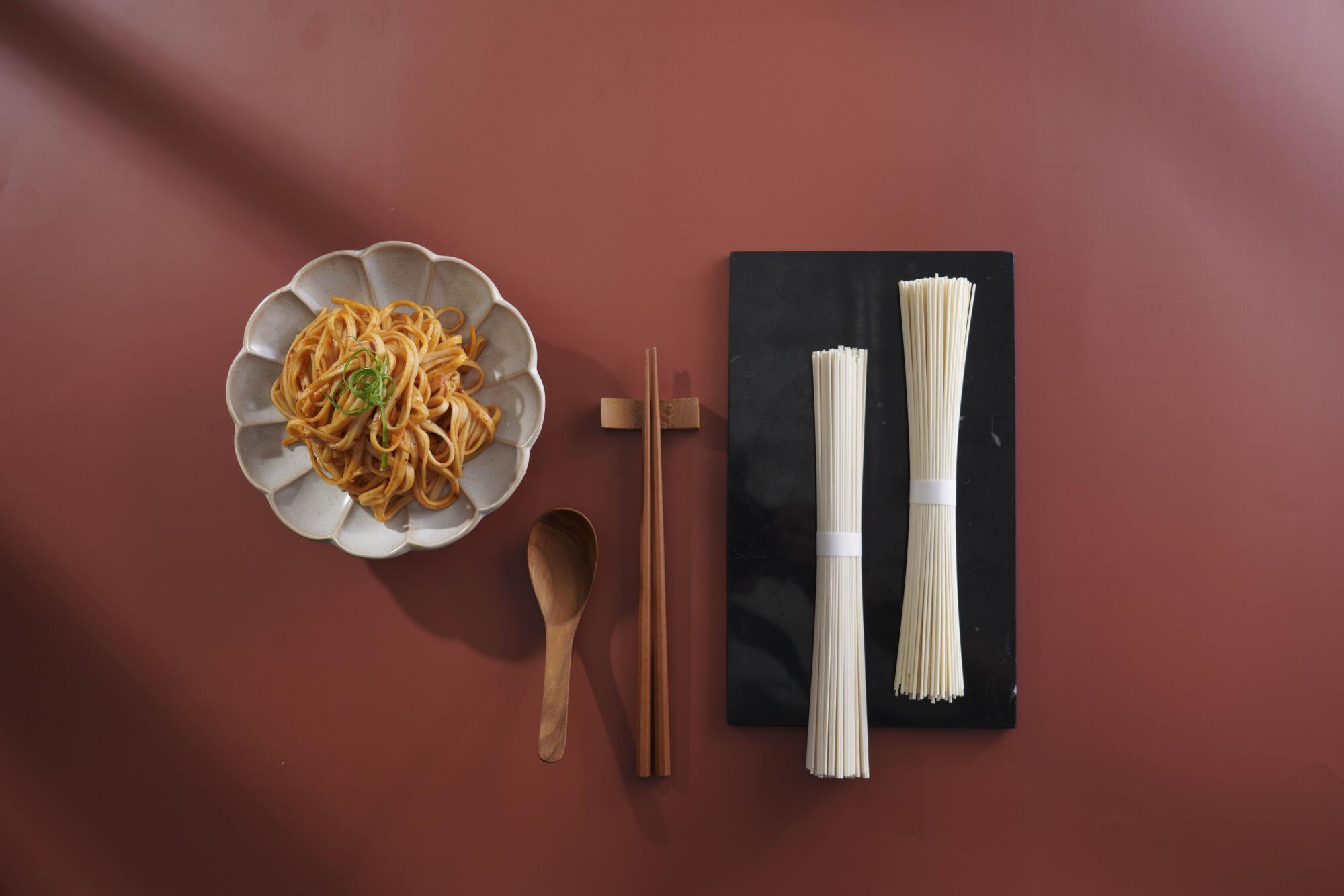 A finished noodle dish displayed beside uncooked noodles of different thicknesses and textures.