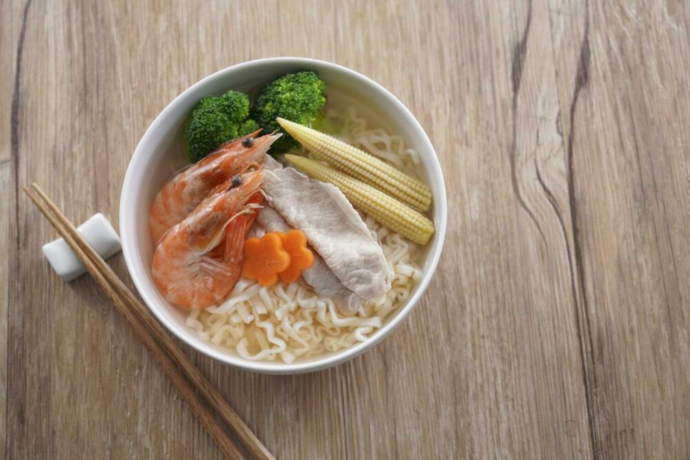A bowl of SING-LIN FOODS ramen noodles served with shrimp, pork slices, broccoli, and baby corn.