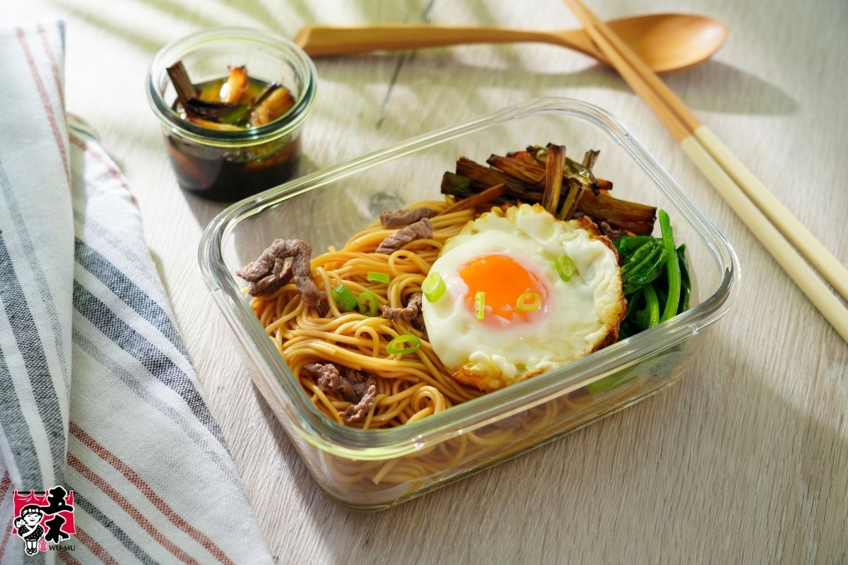 Shanghai scallion oil noodles in a glass lunch container with a fried egg, sliced meat, greens, and browned scallions.