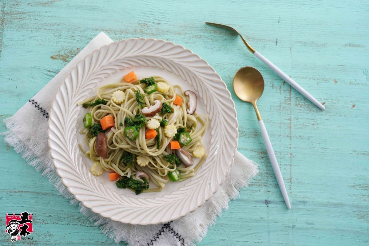 Vegetable noodles with carrots, baby corn, okra, shiitake mushrooms, and leafy greens on a white plate.