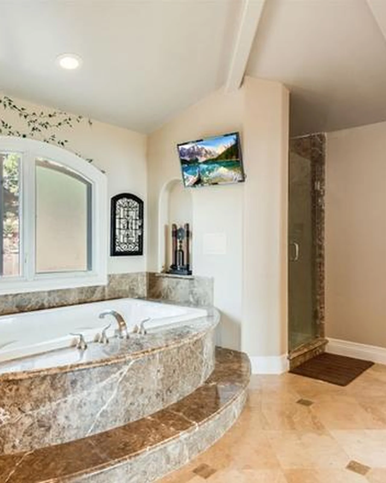 Original primary bathroom with an oval drop-in tub in a travertine surround, brass fixtures, a wall-mounted television and a beige tile floor