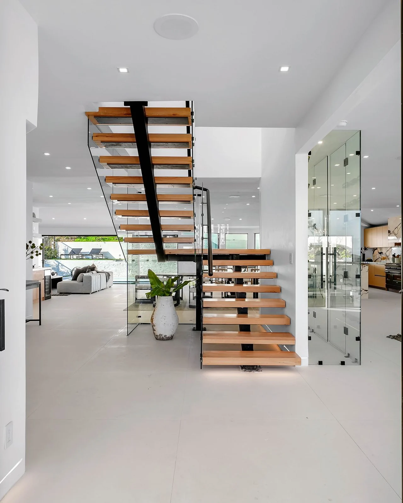 The rebuilt entry, sightline running to an oak flight with open treads carried on darkened steel, guarded in glass with no posts, the pale floor continuing beneath it