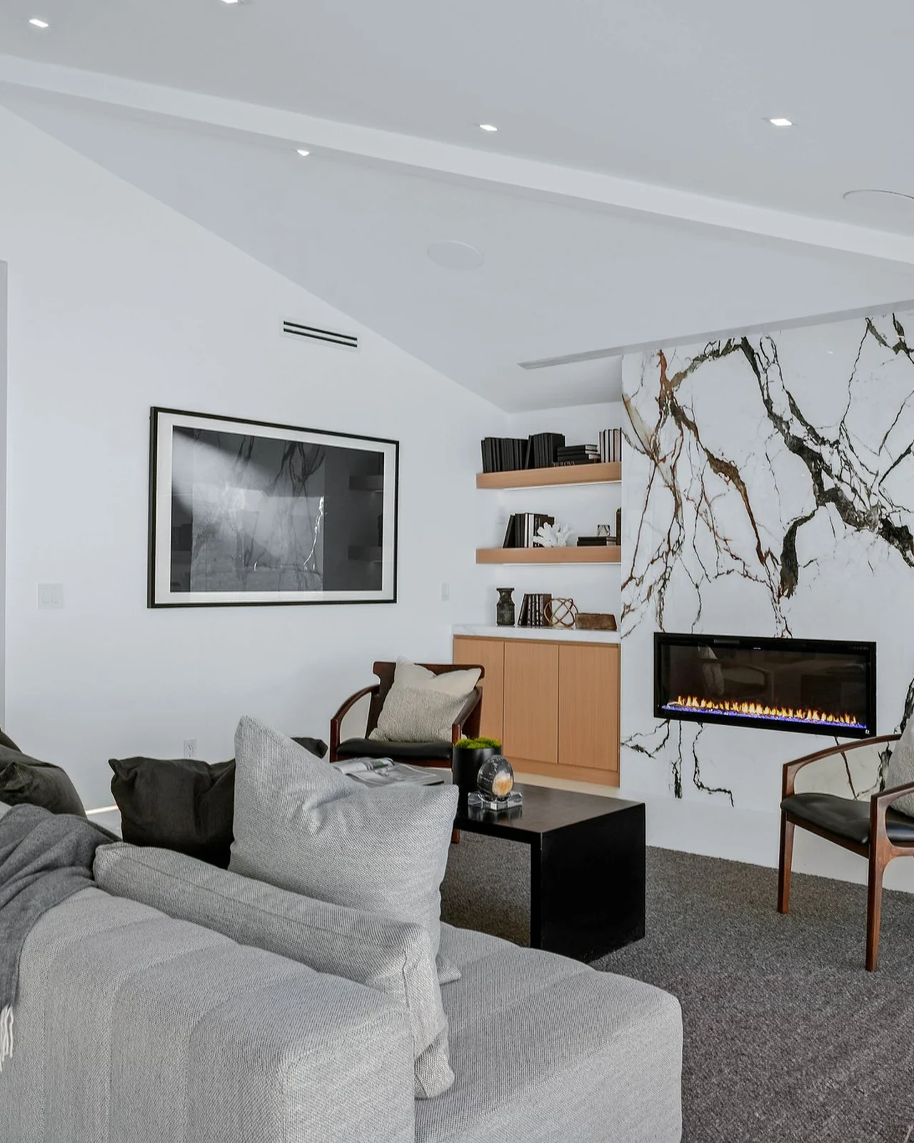 The same room in white with a veined marble slab wall, a linear gas fireplace, floating oak shelving, oversized black-and-white artwork and a low grey sofa