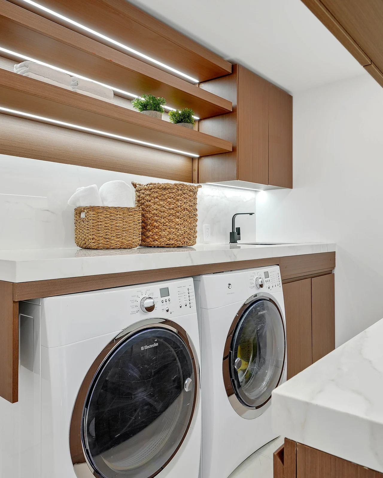 Rebuilt laundry room with walnut-toned cabinetry, integrated LED shelf lighting, a marble counter and backsplash, and machines set under the run