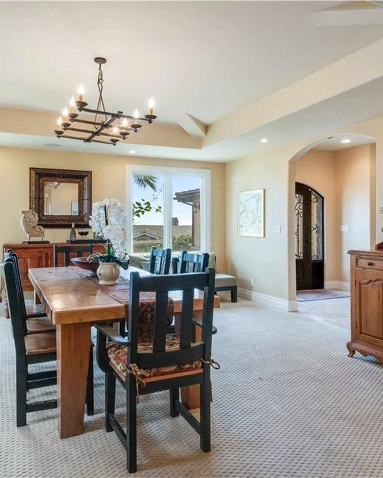 Original formal dining and living room with beige walls, a chunky rustic wood table, an iron chandelier, a framed mirror, carpet and a sightline to the arched front door
