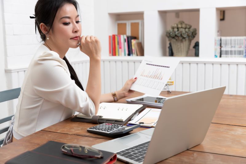Empresária, pensativa, segura um relatório em uma das mãos enquanto analisa as contas a pagar e receber no laptop.