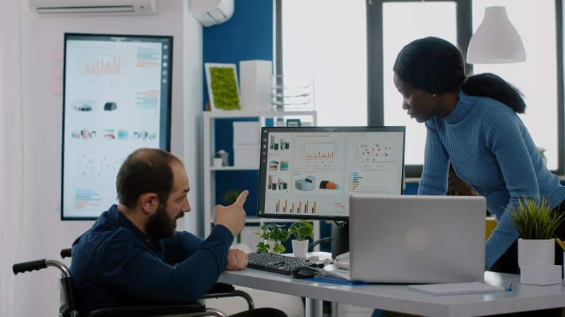 Homem em cadeira de rodas apontando para a tela do computador explica os tipos de métricas para empresas de TI a uma colega que está em pé do outro lado da mesa.