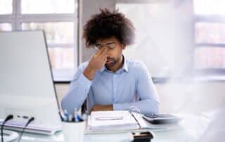 Gestor contábil sentado em frente ao computador e com a mão no rosto, preocupado, pensando como diminuir a inadimplência.