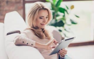 Mulher usando tablet sentada em sofá, lendo com tranquilidade uma notificação de cobrança que recebeu.