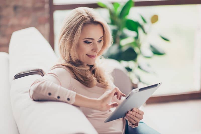 Mulher usando tablet sentada em sofá, lendo com tranquilidade uma notificação de cobrança que recebeu.