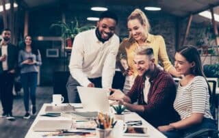 Como funcionam as startups: grupo de mulheres e homens sorridentes reunidos em volta de mesa com computador.