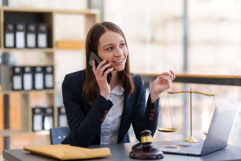 advogada falando com cliente no celular enquanto trabalha em sua mesa, com computador e decorações relacionadas à advocacia