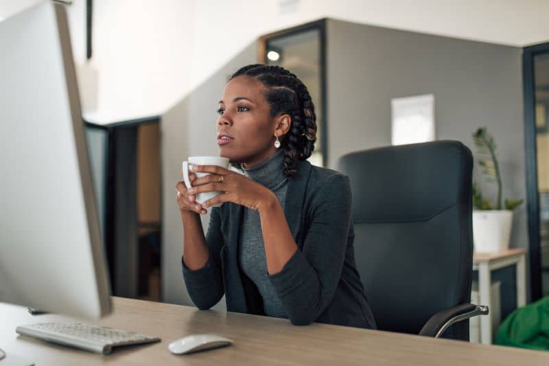 Empresária em seu escritório visualizando relatórios financeiros no computador enquanto segura uma caneca entre as mãos.