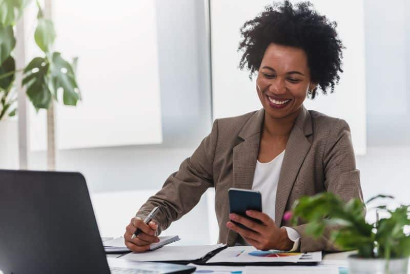 Mulher sorridente em escritório falando com cliente pelo celular sobre reajuste de preços.