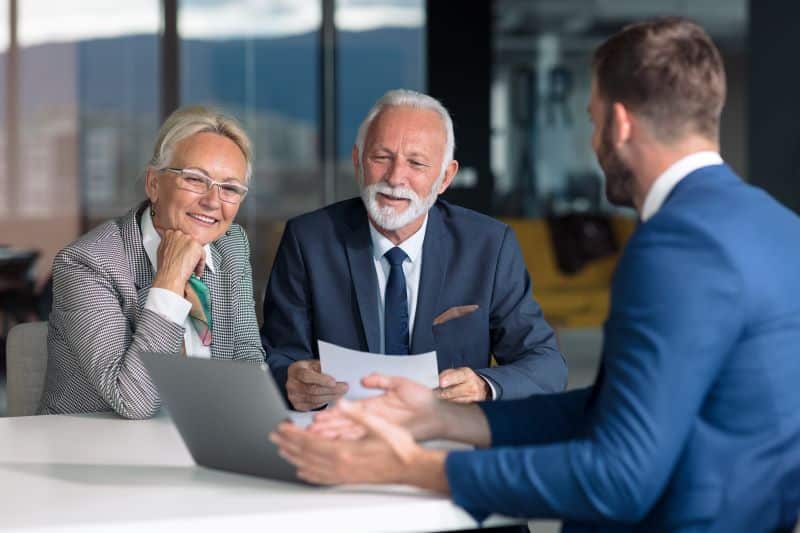 Vendedor apresenta sistema SaaS em notebook para dois clientes sorridentes sentado à sua frente, todos vestidos formalmente.