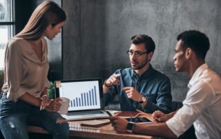 Dashboard o que é​: três profissionais reunidos em volta de mesa analisando gráfico no computador.