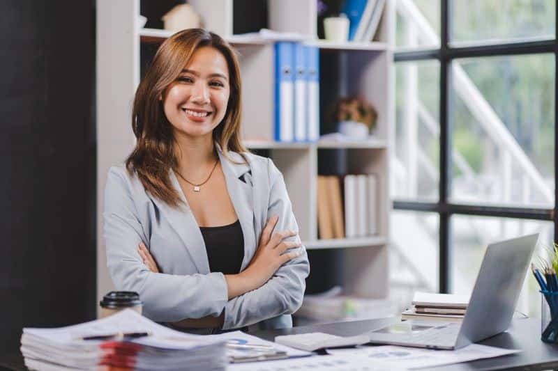 Mulher de braços cruzados sorri para câmera em seu escritório, com documentos referentes a ativos e passivos financeiros sobre a mesa.