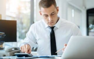 Homem de camisa branca e gravata usando calculadora para verificar taxas, em mesa com computador e telefone.