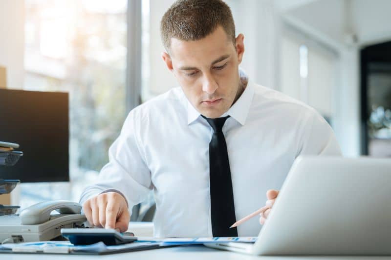 Homem de camisa branca e gravata usando calculadora para verificar taxas, em mesa com computador e telefone.