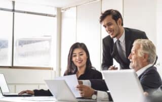 Uma mulher e dois homens, todos vestidos formalmente, em sala de reunião com computadores, analisam contrato SaaS e conversam sobre sua elaboração.