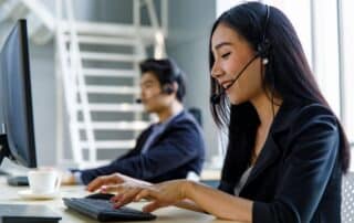 mulher com headphone atendendo ligação de cliente enquanto trabalha no computador, com colega de trabalho ao fundo na mesma função.