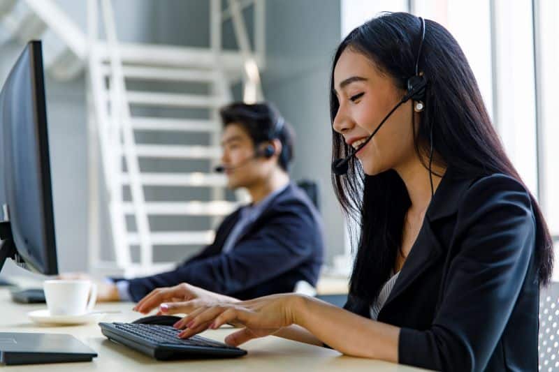 mulher com headphone atendendo ligação de cliente enquanto trabalha no computador, com colega de trabalho ao fundo na mesma função.