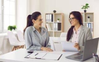 Duas gestoras conversam sobre a previsão financeira da empresa sentadas à mesa com computador e documentos em mãos.