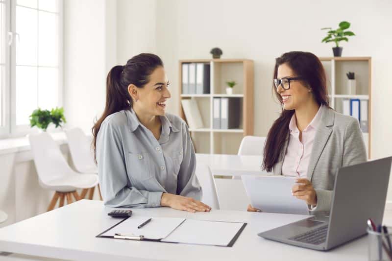 Duas gestoras conversam sobre a previsão financeira da empresa sentadas à mesa com computador e documentos em mãos.