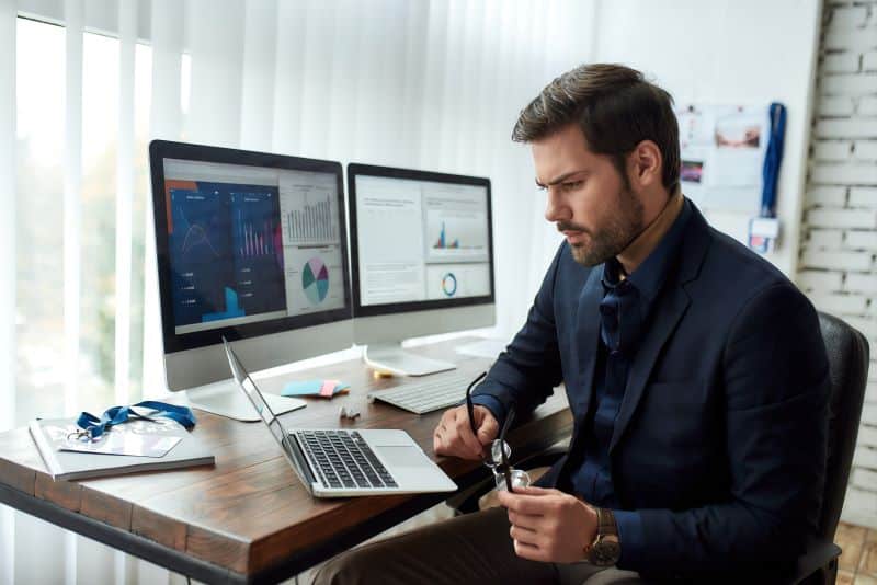 Analista financeiro em seu escritório visualizando dashboards financeiros em notebook, com outros dois monitores ao lado na mesma mesa.