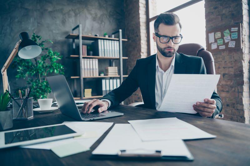 homem de óculos e roupa social sentado em seu escritório trabalhando enquanto analisa relatórios financeiros impressos e eletrônicos