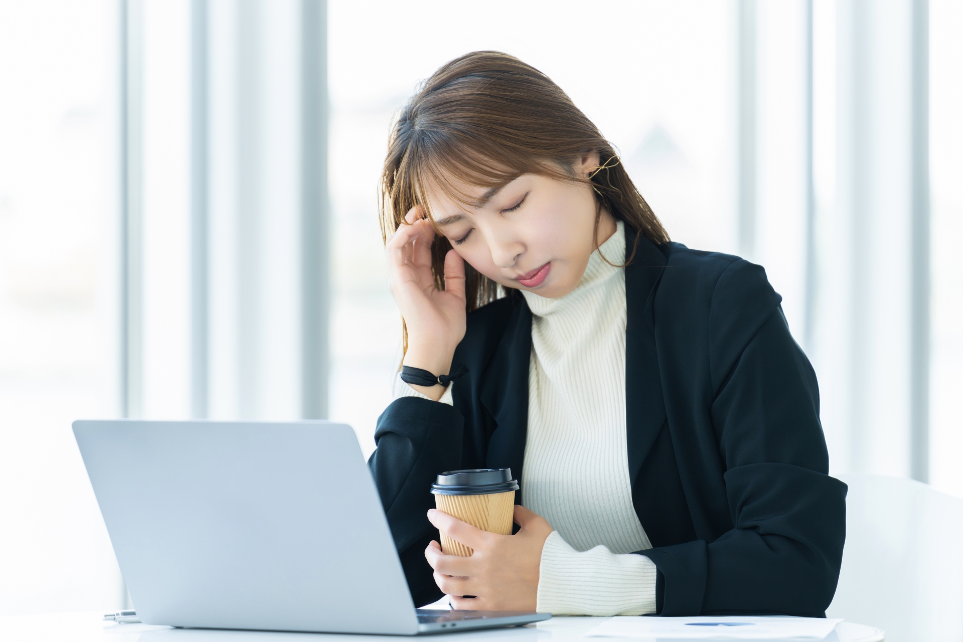 黒いジャケットを着た女性が、ノートPCの前でコーヒーカップを抱えながら、片手でこめかみを押さえて目を閉じている。ヒューリスティック分析のデメリットに直面している。