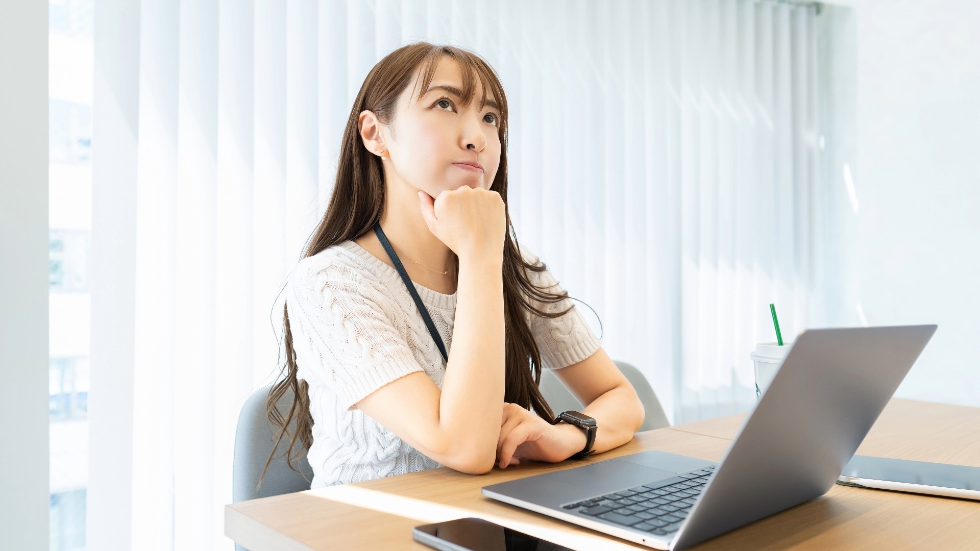 ノートPCが置かれたテーブルで、白い服を着た女性が顎に手を当てて斜め上を見つめ、考え込んでいる表情。ヒューリスティック分析が自社のような小規模サイトでも効果があるのか疑問に感じている。