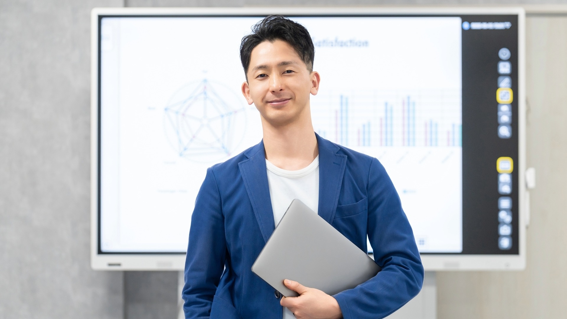 実践的なLLMO・AIO対策を提案する、分析データが映るモニターの前に立ちノートPCを持つ男性コンサルタント。